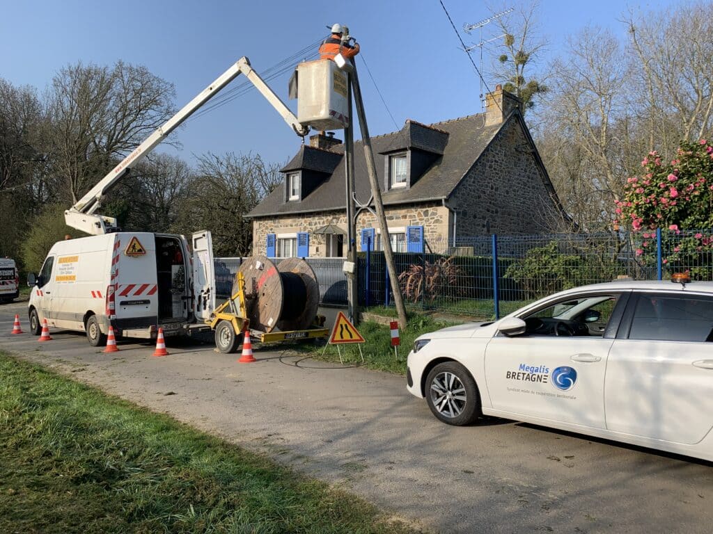 Travaux de déploiement de la fibre optique avec des ouvriers présents sur une nacelle. A côté de celle-ci se trouve une voiture de Mégalis Bretagne